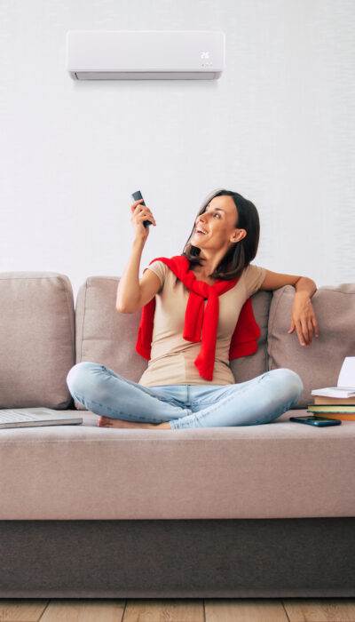 Beautiful modern brunette woman is using the air conditioner while sitting on the couch and resting in the hot day