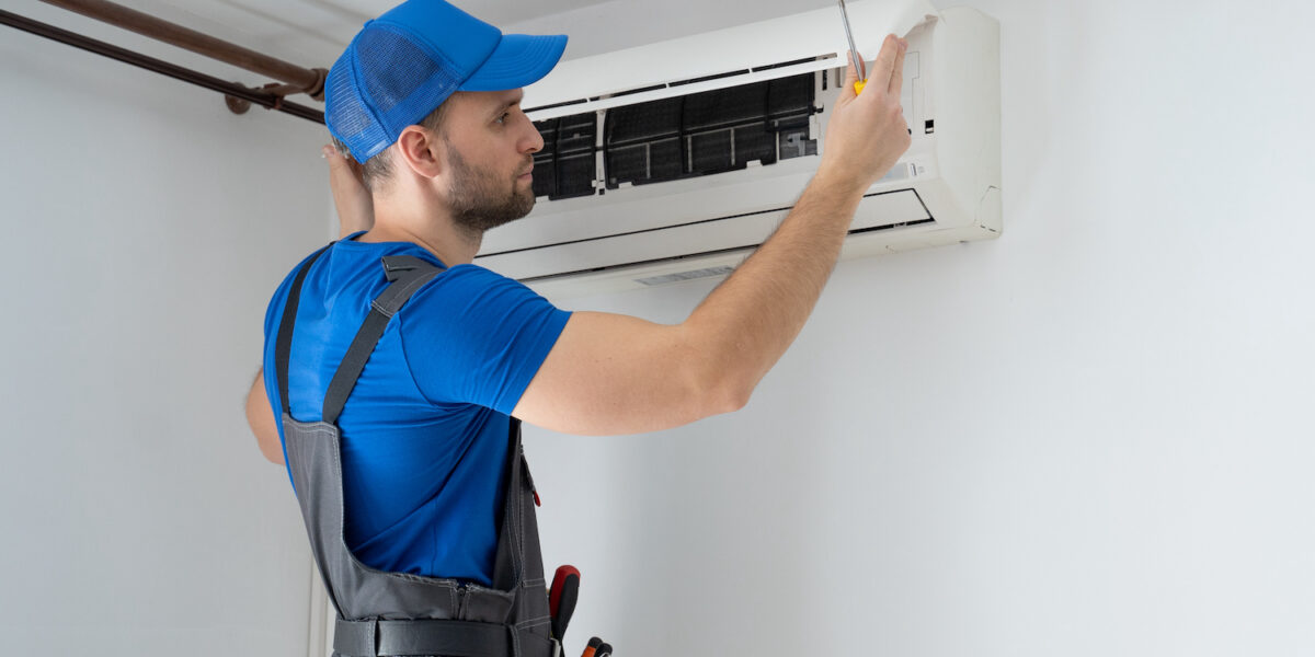 Male technician in overalls and a blue cap repairs an air conditioner on the wall.