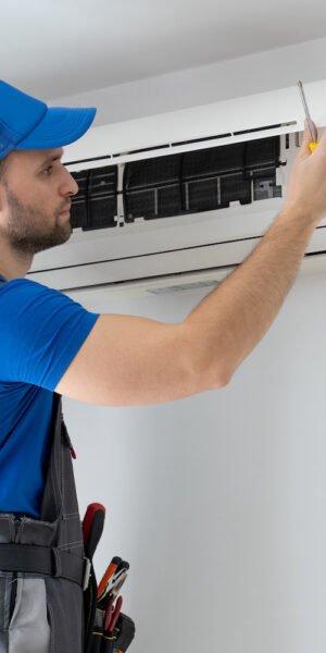 Male technician in overalls and a blue cap repairs an air conditioner on the wall.